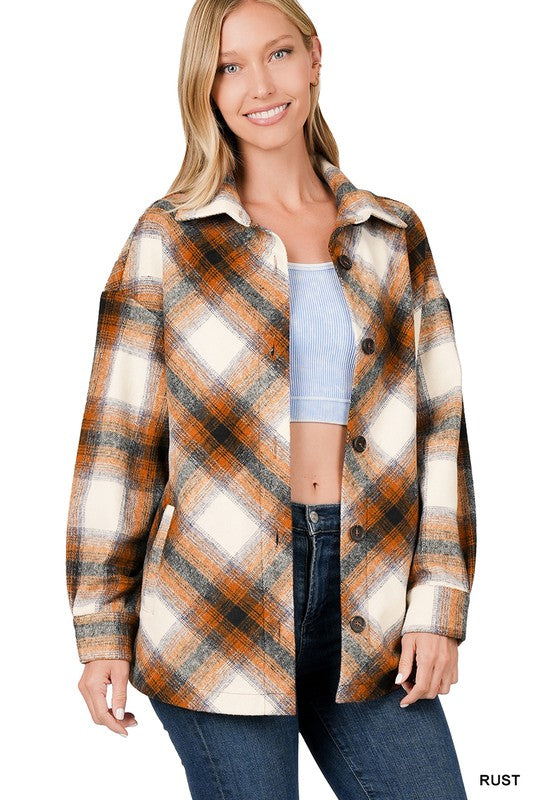 A woman is wearing a Yarn Dyed Plaid Shacket With Pockets in a rust color. One hand is on her collar while the other rests in her pocket. She has light hair and is looking forward.