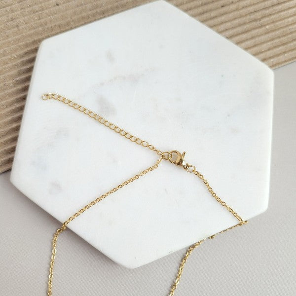 The Ava Blonde Tortoise Necklace is a dainty gold chain necklace with an 18K gold-plated pendant featuring a small gold triangle and a larger speckled half-circle, elegantly displayed on a white hexagonal surface.