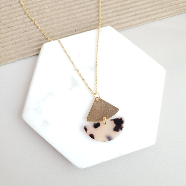 The Ava Blonde Tortoise Necklace is a dainty gold chain necklace with an 18K gold-plated pendant featuring a small gold triangle and a larger speckled half-circle, elegantly displayed on a white hexagonal surface.