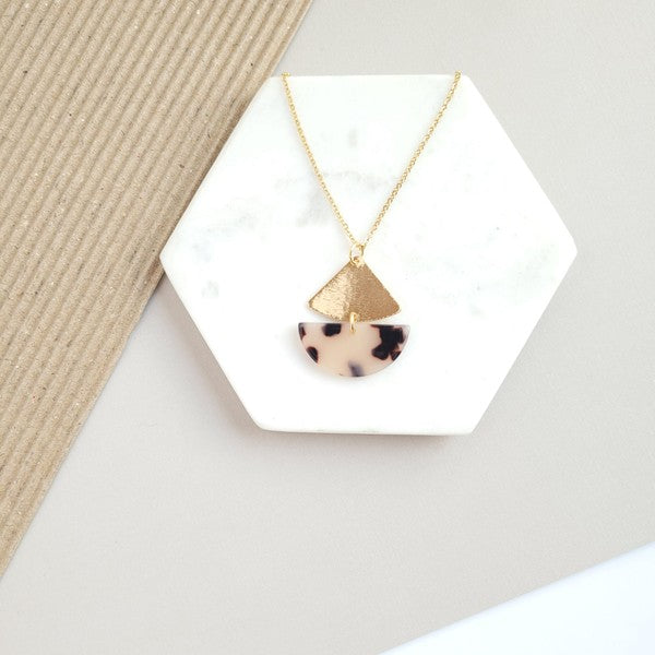 The Ava Blonde Tortoise Necklace is a dainty gold chain necklace with an 18K gold-plated pendant featuring a small gold triangle and a larger speckled half-circle, elegantly displayed on a white hexagonal surface.