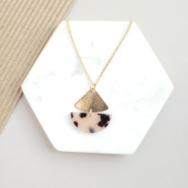 The Ava Blonde Tortoise Necklace is a dainty gold chain necklace with an 18K gold-plated pendant featuring a small gold triangle and a larger speckled half-circle, elegantly displayed on a white hexagonal surface.