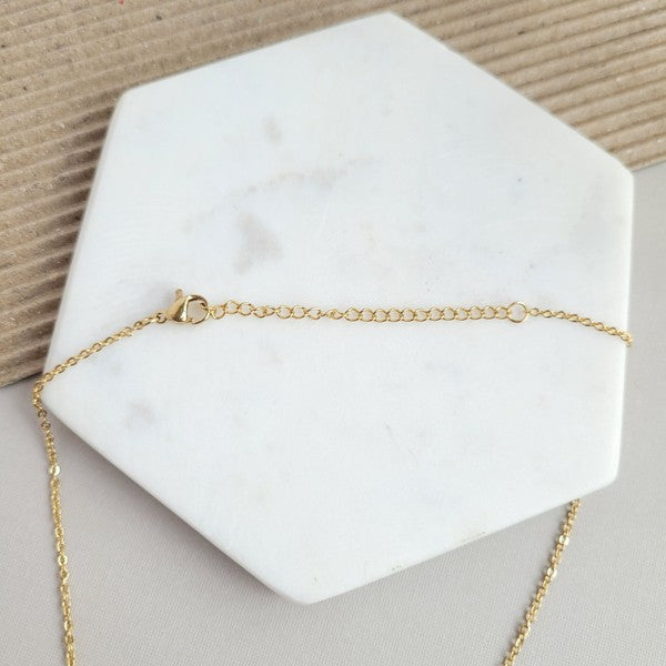 The Ava Blonde Tortoise Necklace is a dainty gold chain necklace with an 18K gold-plated pendant featuring a small gold triangle and a larger speckled half-circle, elegantly displayed on a white hexagonal surface.