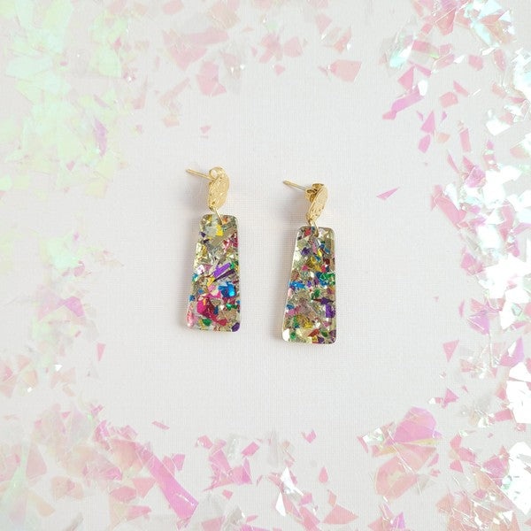 A pair of Mia Mini Earrings - Unicorn, featuring rectangular shapes and multicolored glitter with gold stud tops, are displayed on a white hexagonal dish, surrounded by iridescent confetti.