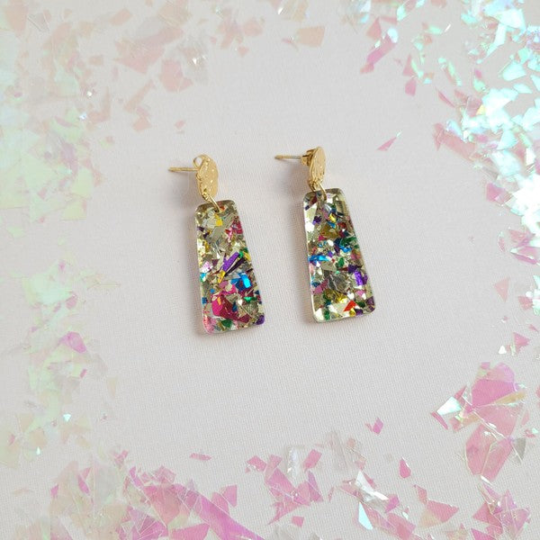 A pair of Mia Mini Earrings - Unicorn, featuring rectangular shapes and multicolored glitter with gold stud tops, are displayed on a white hexagonal dish, surrounded by iridescent confetti.