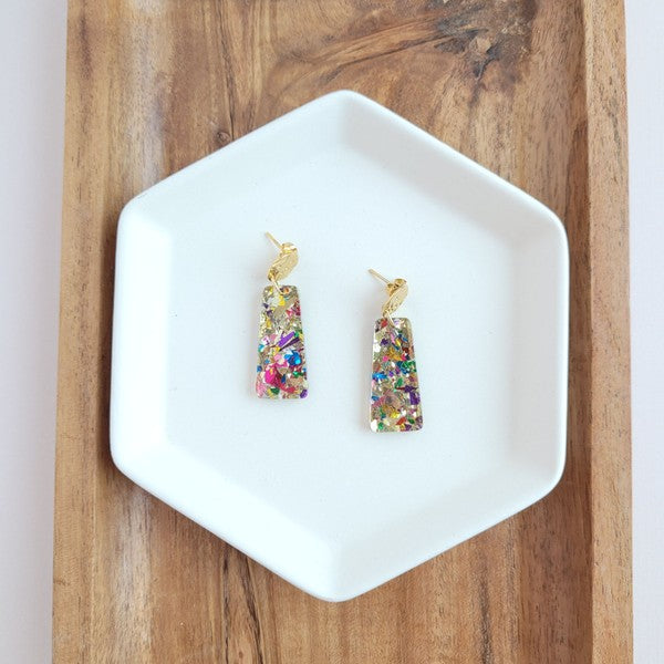 A pair of Mia Mini Earrings - Unicorn, featuring rectangular shapes and multicolored glitter with gold stud tops, are displayed on a white hexagonal dish, surrounded by iridescent confetti.