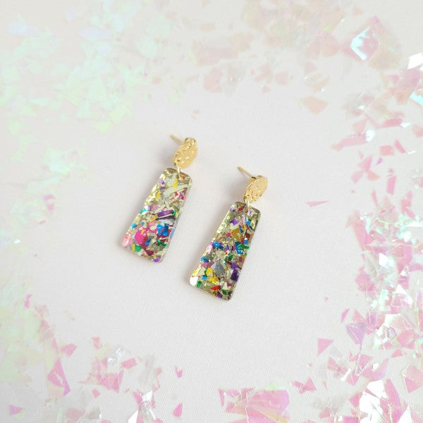 A pair of Mia Mini Earrings - Unicorn, featuring rectangular shapes and multicolored glitter with gold stud tops, are displayed on a white hexagonal dish, surrounded by iridescent confetti.