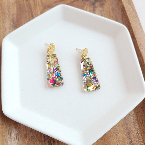 A pair of Mia Mini Earrings - Unicorn, featuring rectangular shapes and multicolored glitter with gold stud tops, are displayed on a white hexagonal dish, surrounded by iridescent confetti.