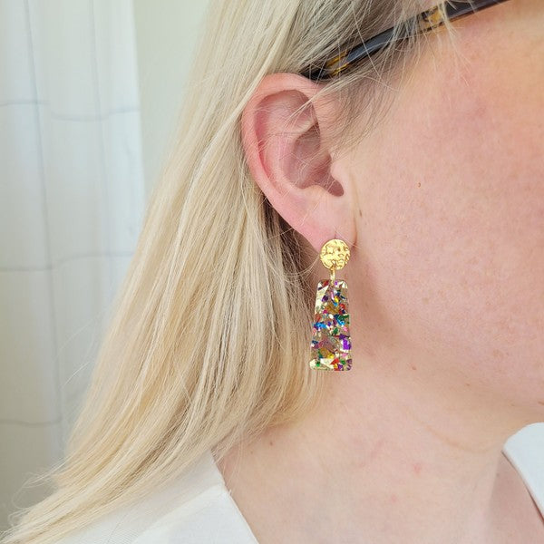 A pair of Mia Mini Earrings - Unicorn, featuring rectangular shapes and multicolored glitter with gold stud tops, are displayed on a white hexagonal dish, surrounded by iridescent confetti.