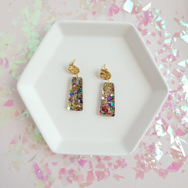 A pair of Mia Mini Earrings - Unicorn, featuring rectangular shapes and multicolored glitter with gold stud tops, are displayed on a white hexagonal dish, surrounded by iridescent confetti.