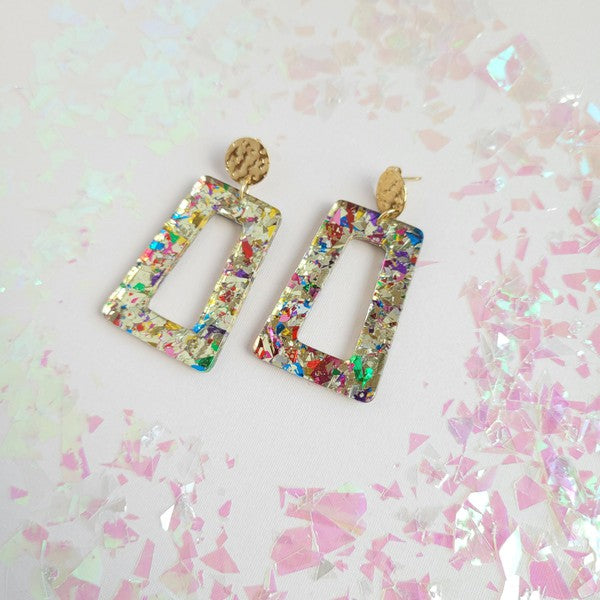 A pair of Avery - Unicorn earrings, rectangular and multi-colored with glitter, featuring 18K gold-plated studs, displayed on a white hexagonal dish on a wooden surface.