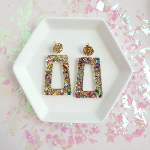 A pair of Avery - Unicorn earrings, rectangular and multi-colored with glitter, featuring 18K gold-plated studs, displayed on a white hexagonal dish on a wooden surface.