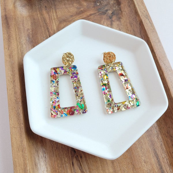 A pair of Avery - Unicorn earrings, rectangular and multi-colored with glitter, featuring 18K gold-plated studs, displayed on a white hexagonal dish on a wooden surface.