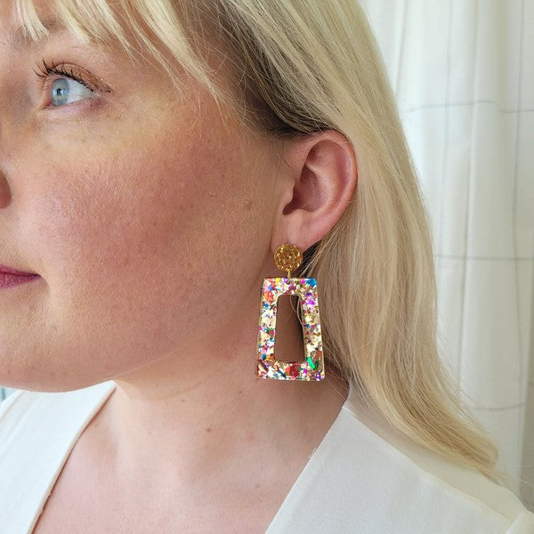 A pair of Avery - Unicorn earrings, rectangular and multi-colored with glitter, featuring 18K gold-plated studs, displayed on a white hexagonal dish on a wooden surface.