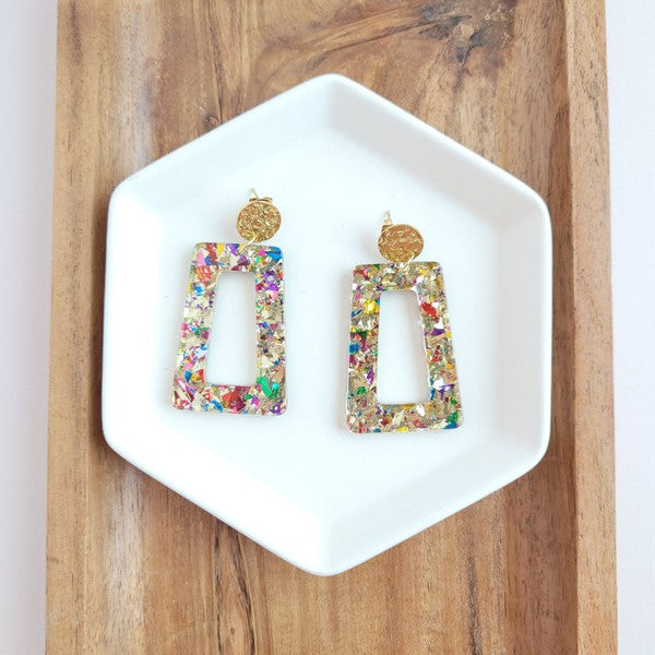 A pair of Avery - Unicorn earrings, rectangular and multi-colored with glitter, featuring 18K gold-plated studs, displayed on a white hexagonal dish on a wooden surface.