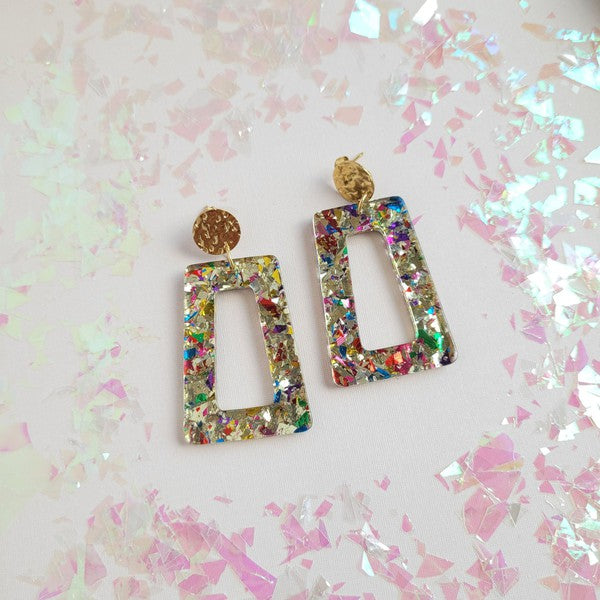 A pair of Avery - Unicorn earrings, rectangular and multi-colored with glitter, featuring 18K gold-plated studs, displayed on a white hexagonal dish on a wooden surface.