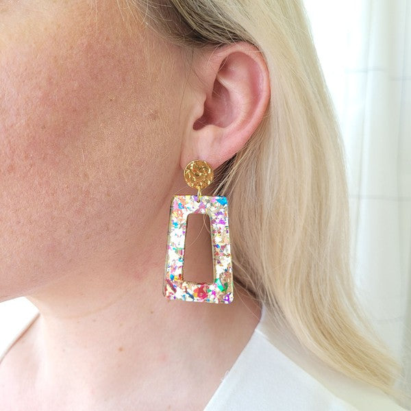 A pair of Avery - Unicorn earrings, rectangular and multi-colored with glitter, featuring 18K gold-plated studs, displayed on a white hexagonal dish on a wooden surface.