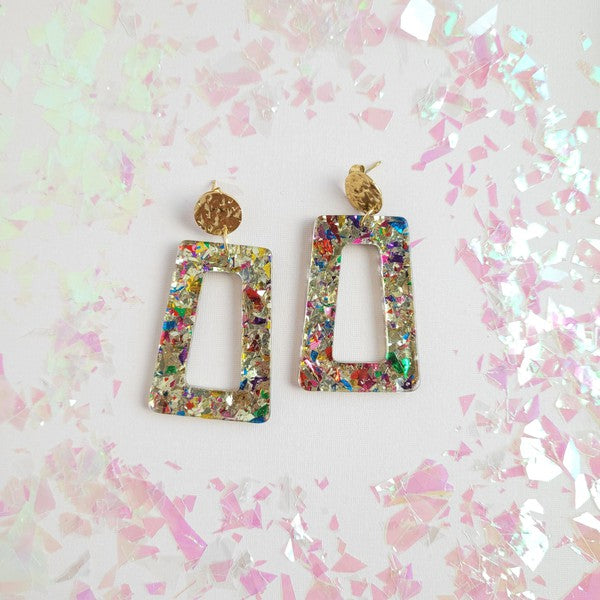 A pair of Avery - Unicorn earrings, rectangular and multi-colored with glitter, featuring 18K gold-plated studs, displayed on a white hexagonal dish on a wooden surface.