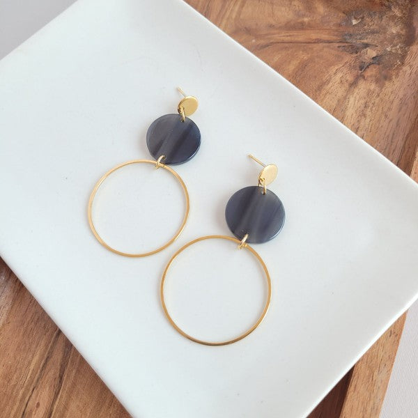 The Sadie Earrings - Black, a pair of gold-plated dangle earrings featuring dark circular acrylic charms, are elegantly displayed on a white rectangular dish set on a wooden surface.