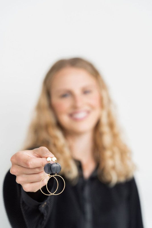 The Sadie Earrings - Black, a pair of gold-plated dangle earrings featuring dark circular acrylic charms, are elegantly displayed on a white rectangular dish set on a wooden surface.