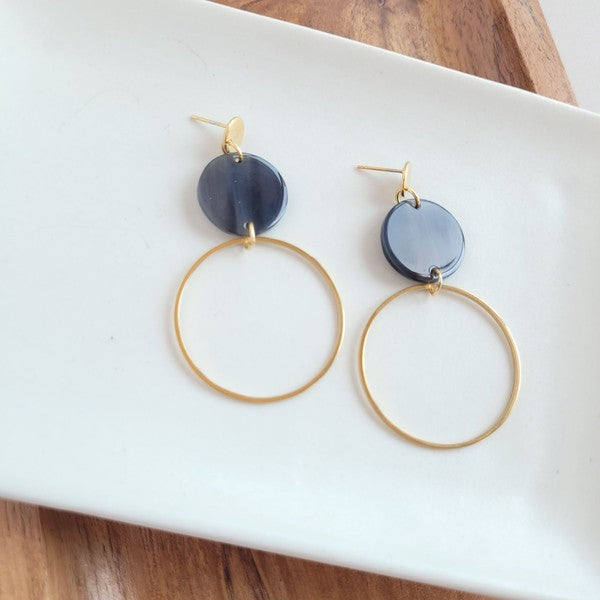 The Sadie Earrings - Black, a pair of gold-plated dangle earrings featuring dark circular acrylic charms, are elegantly displayed on a white rectangular dish set on a wooden surface.