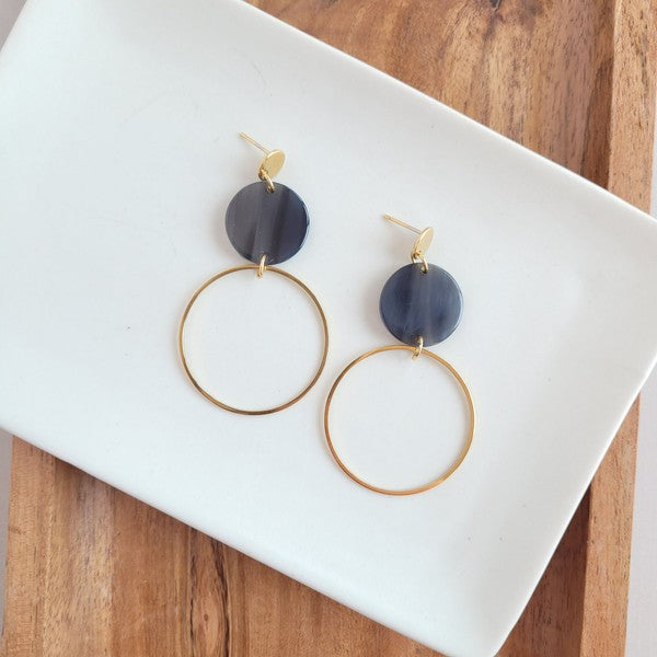 The Sadie Earrings - Black, a pair of gold-plated dangle earrings featuring dark circular acrylic charms, are elegantly displayed on a white rectangular dish set on a wooden surface.