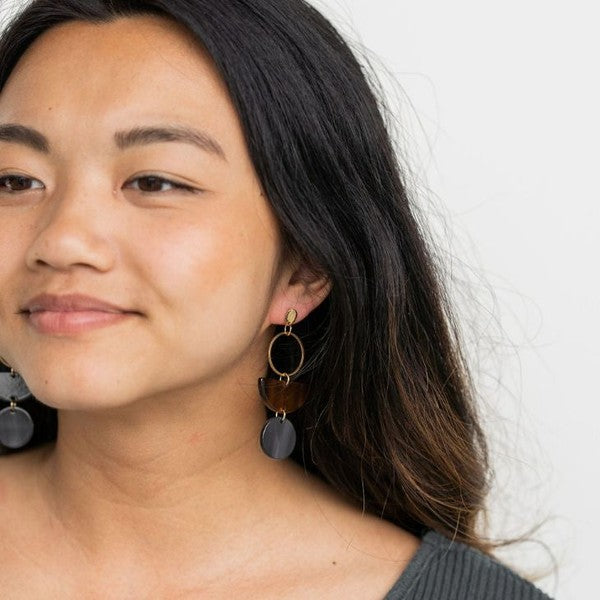 Displayed on a white rectangular dish atop a wooden surface, the Wren Walnut and Black earrings feature a gold hoop, a brown semicircle, and a dark round pendant, crafted from 18k gold-plated stainless steel and lightweight durable acrylic.