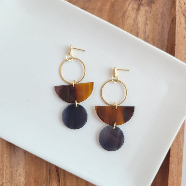 Displayed on a white rectangular dish atop a wooden surface, the Wren Walnut and Black earrings feature a gold hoop, a brown semicircle, and a dark round pendant, crafted from 18k gold-plated stainless steel and lightweight durable acrylic.