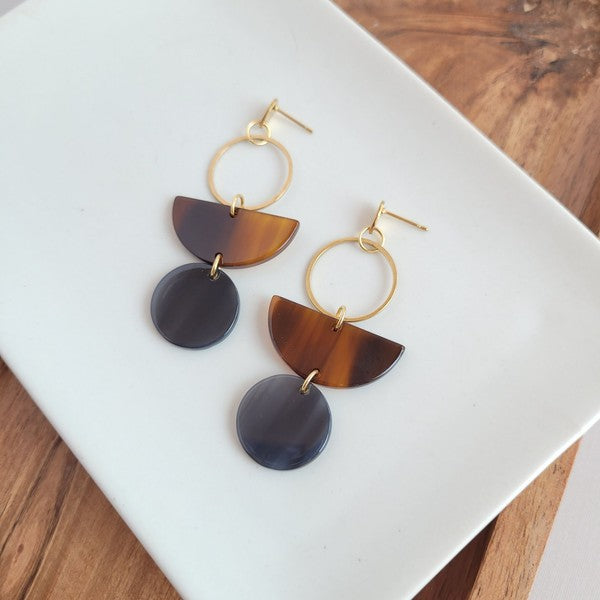 Displayed on a white rectangular dish atop a wooden surface, the Wren Walnut and Black earrings feature a gold hoop, a brown semicircle, and a dark round pendant, crafted from 18k gold-plated stainless steel and lightweight durable acrylic.