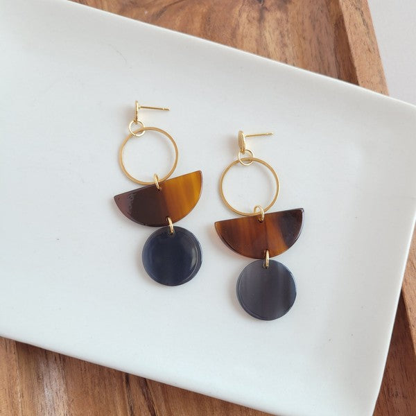 Displayed on a white rectangular dish atop a wooden surface, the Wren Walnut and Black earrings feature a gold hoop, a brown semicircle, and a dark round pendant, crafted from 18k gold-plated stainless steel and lightweight durable acrylic.