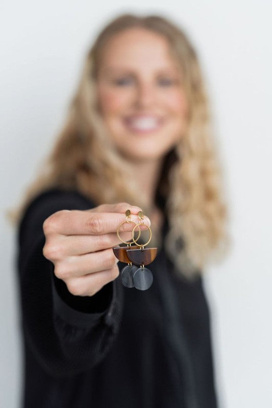 Displayed on a white rectangular dish atop a wooden surface, the Wren Walnut and Black earrings feature a gold hoop, a brown semicircle, and a dark round pendant, crafted from 18k gold-plated stainless steel and lightweight durable acrylic.