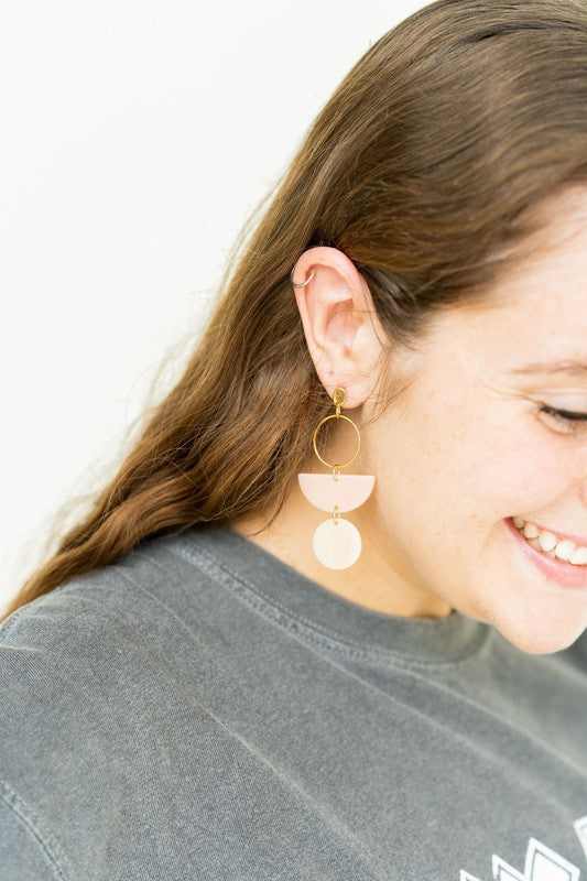 The Wren Blush and Linen statement dangle earrings feature 18k gold-plated rings, pink half-circle acrylic charms, and beige round charms, beautifully displayed on a white rectangular dish with a wooden background.