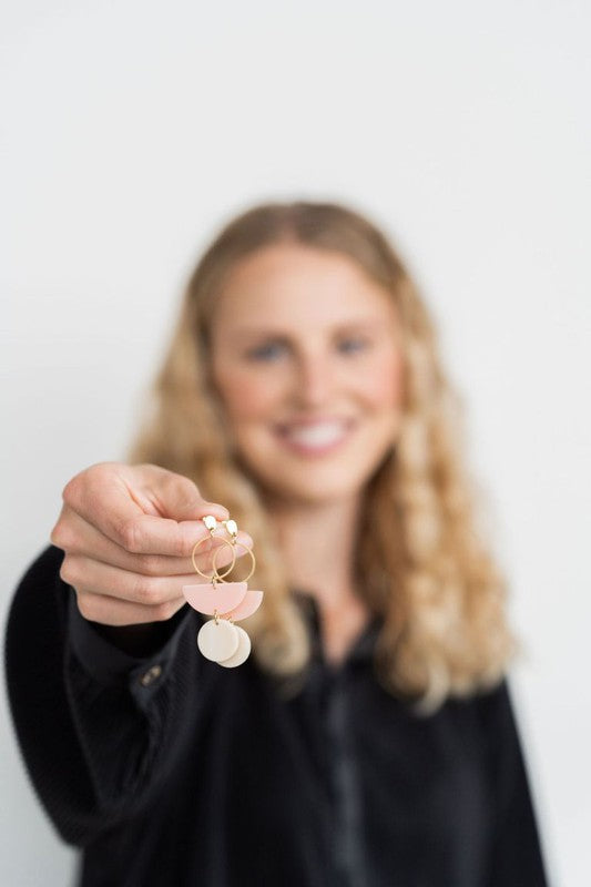 The Wren Blush and Linen statement dangle earrings feature 18k gold-plated rings, pink half-circle acrylic charms, and beige round charms, beautifully displayed on a white rectangular dish with a wooden background.