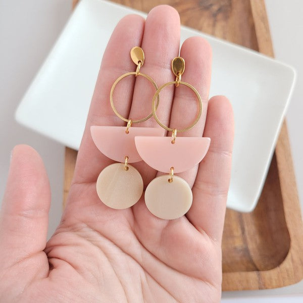 The Wren Blush and Linen statement dangle earrings feature 18k gold-plated rings, pink half-circle acrylic charms, and beige round charms, beautifully displayed on a white rectangular dish with a wooden background.