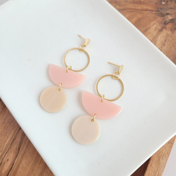 The Wren Blush and Linen statement dangle earrings feature 18k gold-plated rings, pink half-circle acrylic charms, and beige round charms, beautifully displayed on a white rectangular dish with a wooden background.