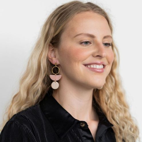 The Wren Blush and Linen statement dangle earrings feature 18k gold-plated rings, pink half-circle acrylic charms, and beige round charms, beautifully displayed on a white rectangular dish with a wooden background.