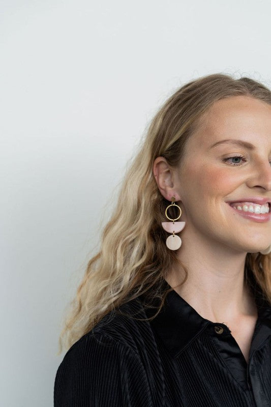 The Wren Blush and Linen statement dangle earrings feature 18k gold-plated rings, pink half-circle acrylic charms, and beige round charms, beautifully displayed on a white rectangular dish with a wooden background.