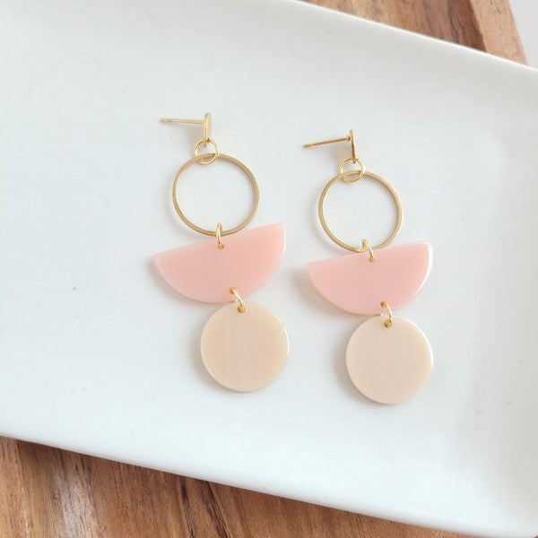 The Wren Blush and Linen statement dangle earrings feature 18k gold-plated rings, pink half-circle acrylic charms, and beige round charms, beautifully displayed on a white rectangular dish with a wooden background.