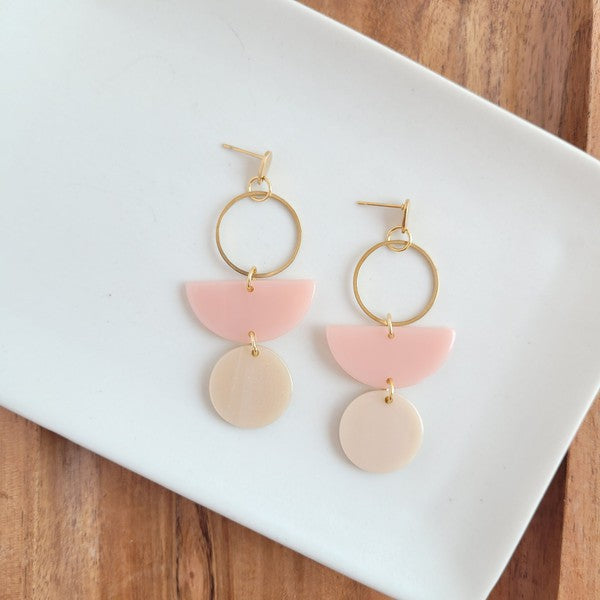 The Wren Blush and Linen statement dangle earrings feature 18k gold-plated rings, pink half-circle acrylic charms, and beige round charms, beautifully displayed on a white rectangular dish with a wooden background.