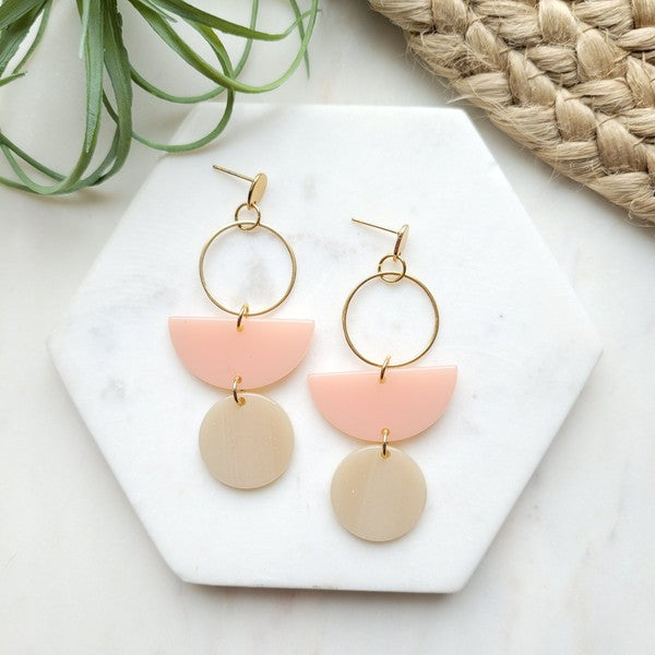 The Wren Blush and Linen statement dangle earrings feature 18k gold-plated rings, pink half-circle acrylic charms, and beige round charms, beautifully displayed on a white rectangular dish with a wooden background.