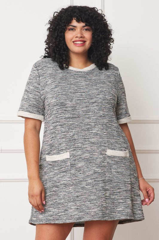 A woman with curly hair is standing in front of a white wall, smiling in a Plus Preppy Tweed Shift Dress featuring a grey design with white trim and two front pockets.