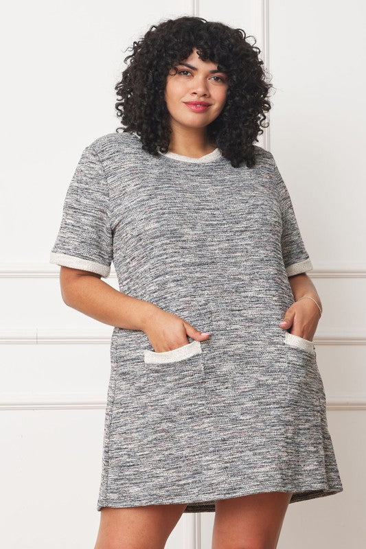 A woman with curly hair is wearing a Plus Preppy Tweed Shift Dress, a textured gray ensemble with white trim and pockets, made from cotton polyester. She stands against a white wall.