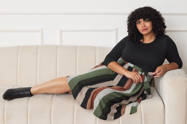 A person stands smiling, wearing a Plus Quarter Sleeve, Stripe Sash Midi Dress. The background is a white wall with decorative molding.