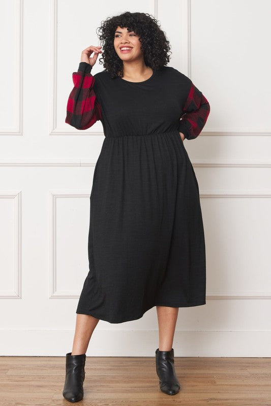 A person stands smiling, wearing the Knit Bishop Sleeve Tea Length Dress with red checkered sleeves and black ankle boots. They have curly hair and are posing with one hand on their head in front of a white paneled wall.