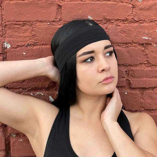 Woman with a pink Cardio Cross-Training Headband made of high-performance elastic fabric, standing against a black brick wall.