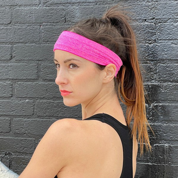 Woman with a pink Cardio Cross-Training Headband made of high-performance elastic fabric, standing against a black brick wall.