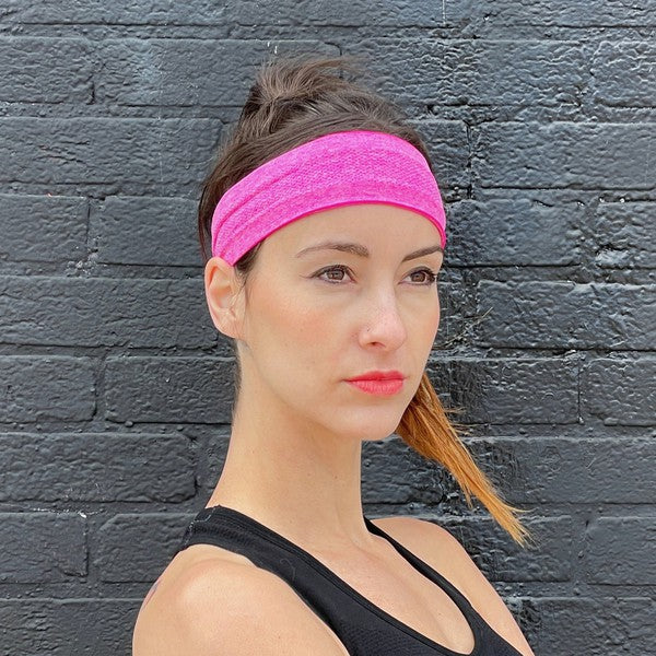 Woman with a pink Cardio Cross-Training Headband made of high-performance elastic fabric, standing against a black brick wall.