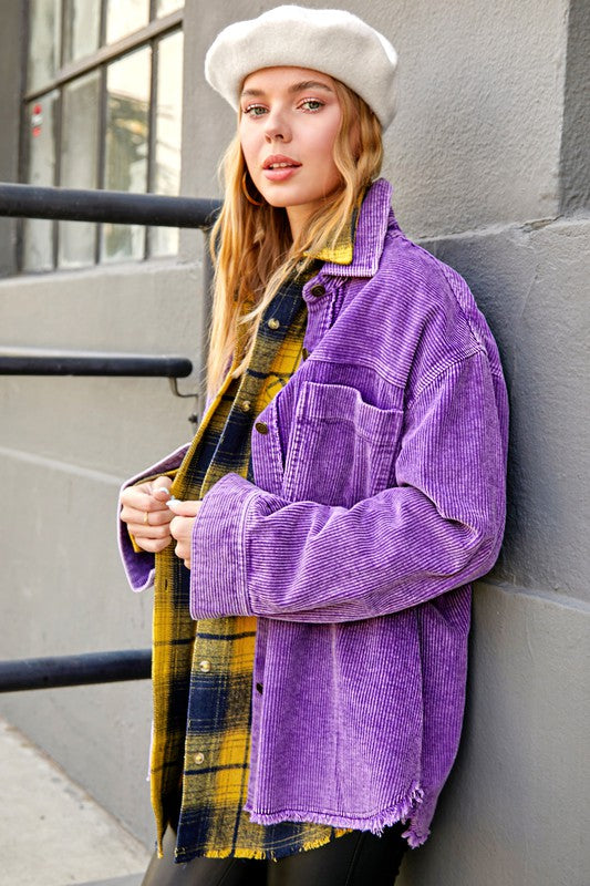 A smiling person wearing sunglasses, the Daisy Jacket in an oversized purple corduroy with a washed finish, and a layered plaid shirt stands outdoors by a light green wall.