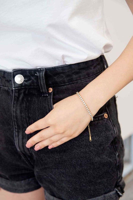 The Round Stone Tennis Bracelet is a silver chain bracelet adorned with clear, round gemstones arranged in a line. It features an adjustable clasp and is plated with 14k gold, presented on a white background.