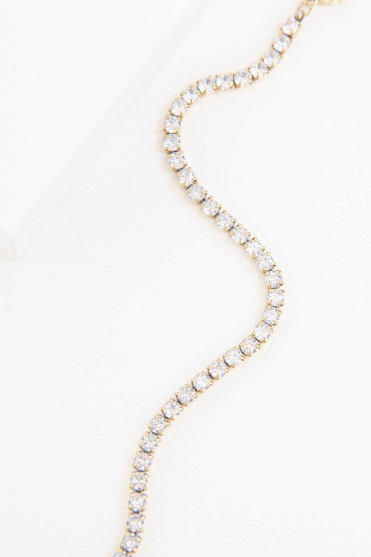 The Round Stone Tennis Bracelet is a silver chain bracelet adorned with clear, round gemstones arranged in a line. It features an adjustable clasp and is plated with 14k gold, presented on a white background.