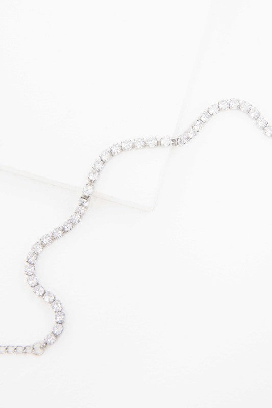 The Round Stone Tennis Bracelet is a silver chain bracelet adorned with clear, round gemstones arranged in a line. It features an adjustable clasp and is plated with 14k gold, presented on a white background.